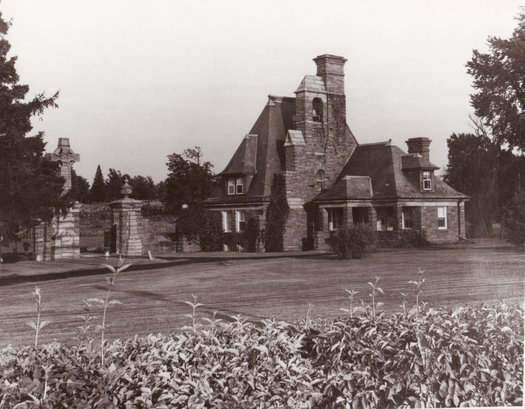 Bayside Lodge, Potsdam, New York (1899). A sepia photograph of a large, angular sandstone building in front of a cemetary, surrounded by black trees.