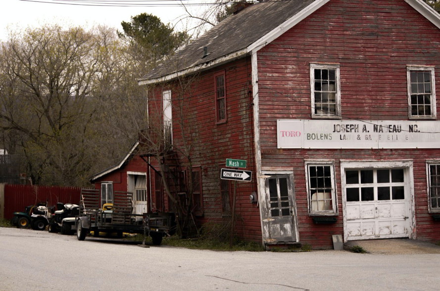 Mower Shop, North Bennington, Vermont (2016). A building faded red siding and weathered windowframes leans noticeably to the left. Several riding mowers and a hitch trailer sit beside it. A street sign out front reads, 'Nash St. ONE WAY.'