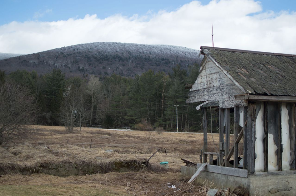 Shaftsbury, Vermont (2017). A white shed against a backdrop of brown fields and snowy mountains. One entire side of the shed has fallen in.